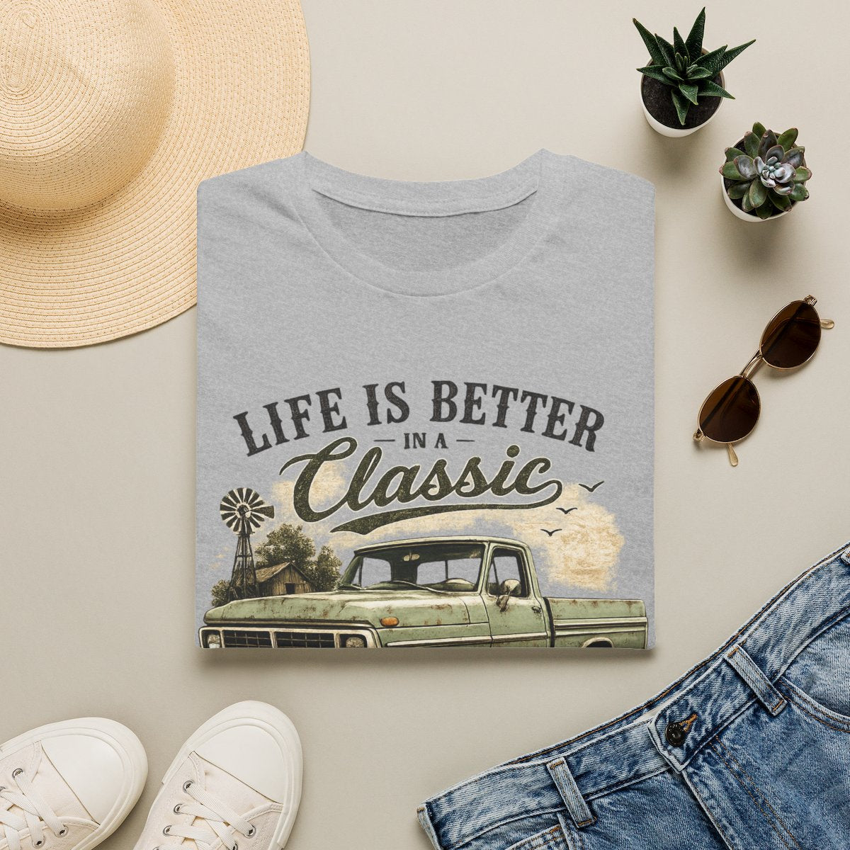 Classic Pickup T-Shirt — "Life Is Better in a Classic" Vintage Truck Tee laid flat on a beige surface, surrounded by a straw hat, white sneakers, blue jeans, sunglasses, and small potted succulents. 