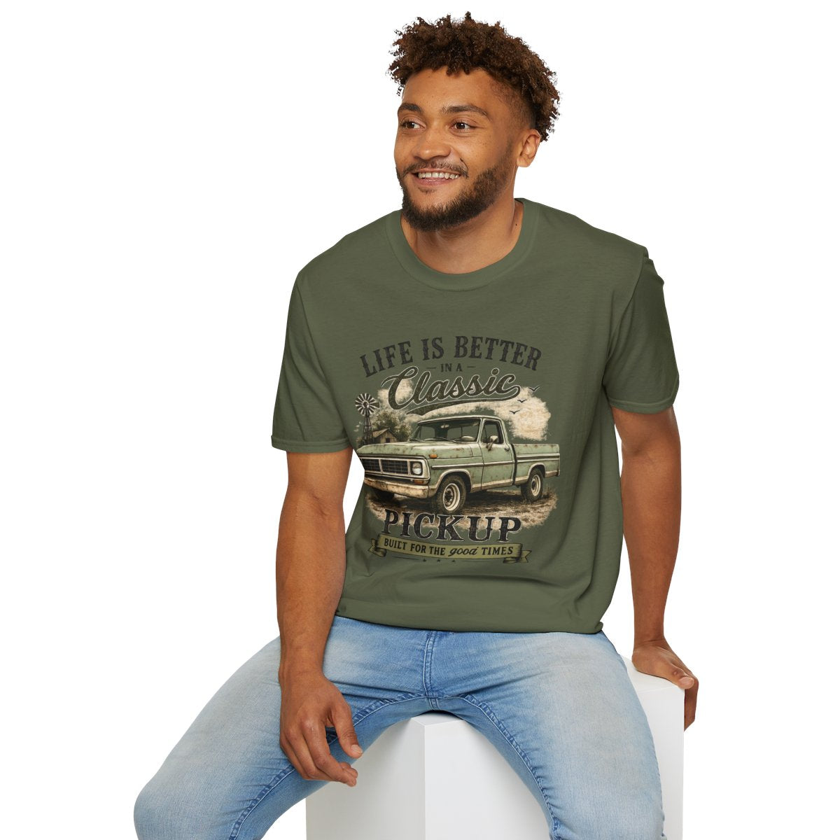 Man wearing Military Green Classic Pickup T-Shirt — "Life Is Better in a Classic" Vintage Truck Tee, sitting on a white stool against a white background.