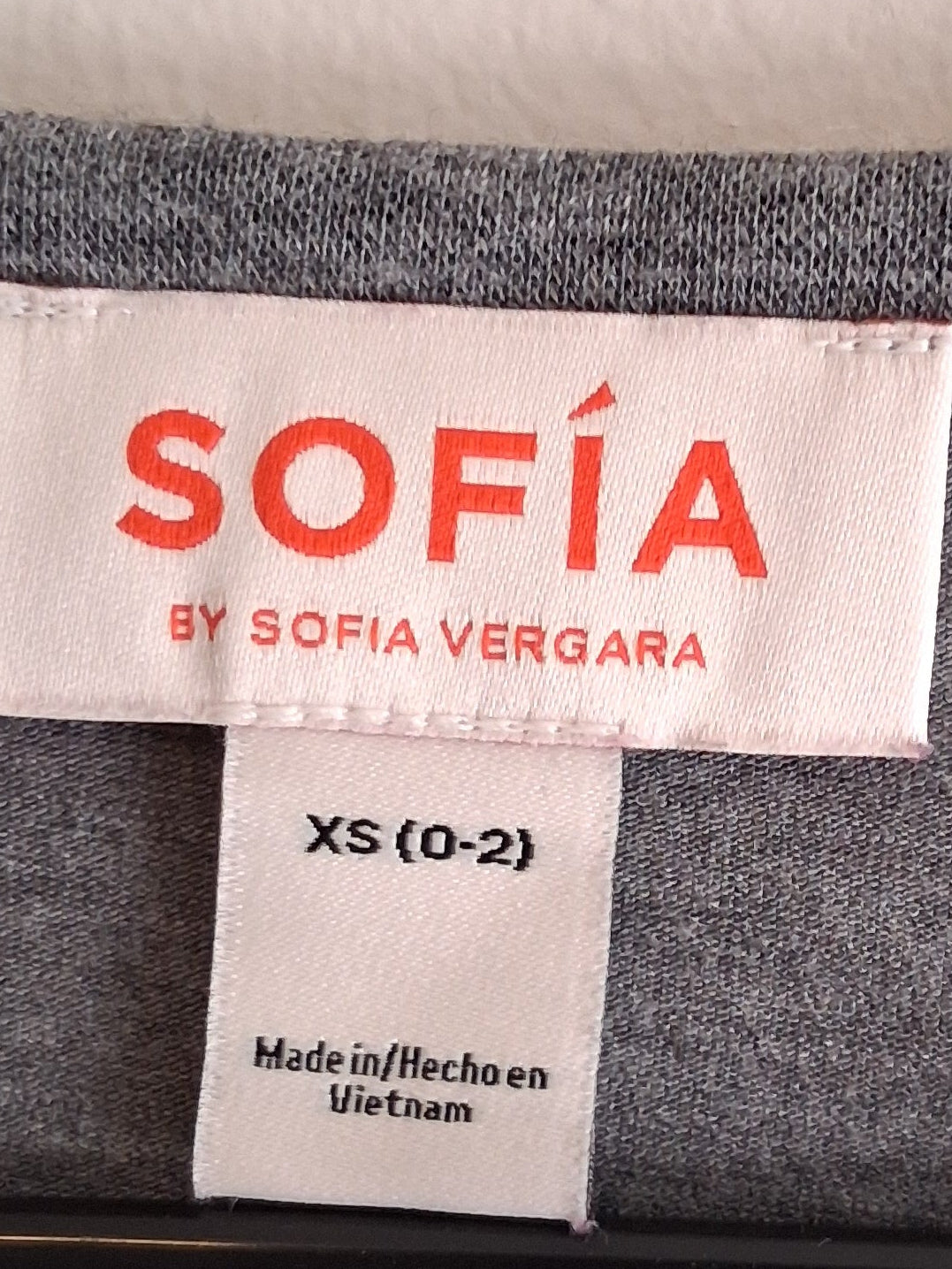 Label with 'SOFIA by Sofia Vergara' text on a gray fabric background - Love is All You Need Tee