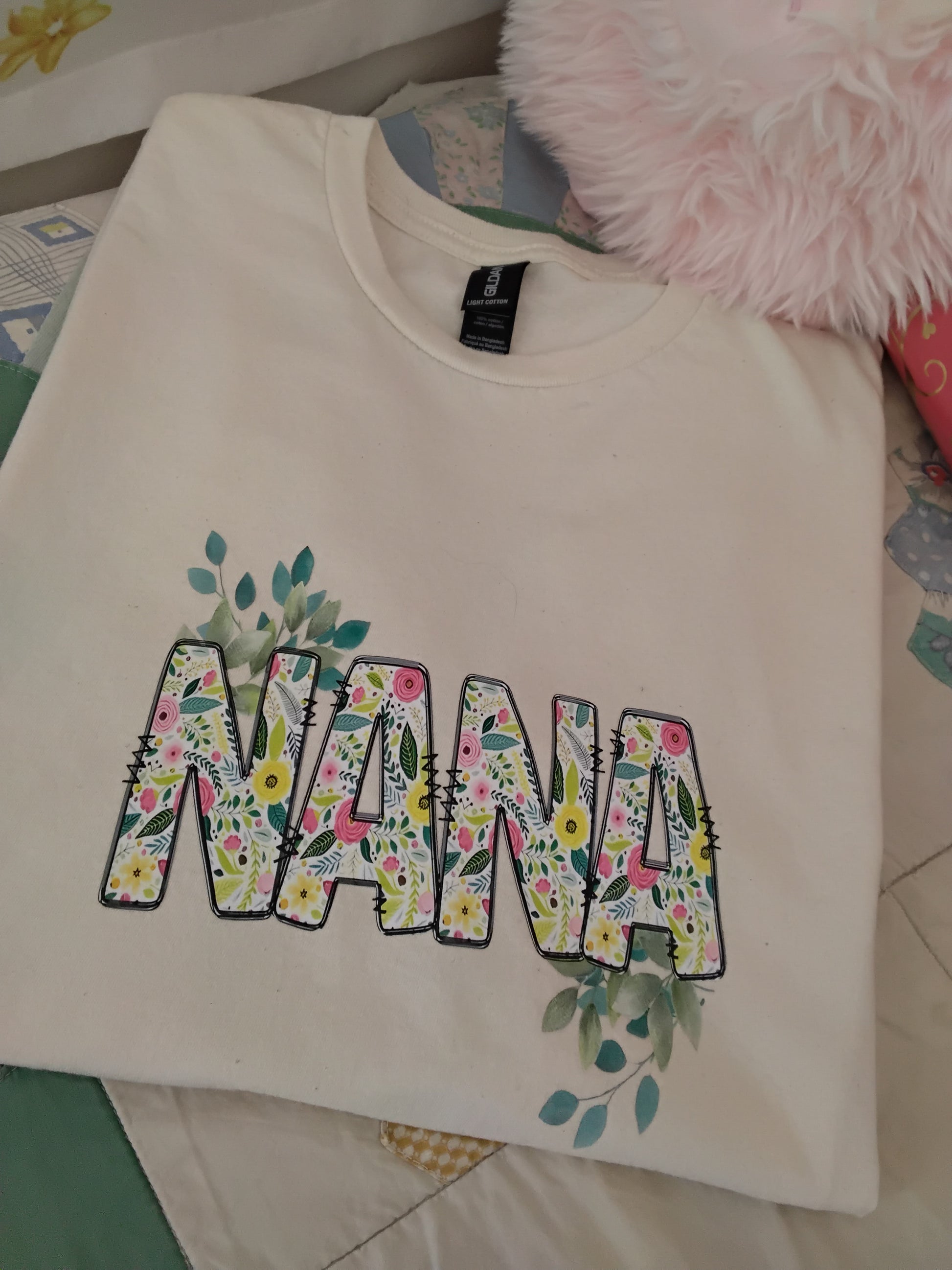 Nana Floral Tee in Natural on a surface with a pink fluffy object and colorful fabric in the background.